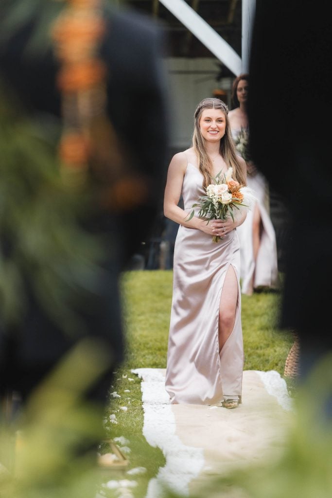 A woman wearing a lightcolored dress walks down an outdoor aisle at The Barns at Hamilton Station, holding a bouquet of flowers, during the wedding ceremony.