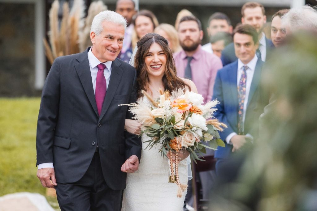 A bride holding a bouquet walks arminarm with a man in a suit down an outdoor aisle at The Barns at Hamilton Station, with guests seated in the background, celebrating this beautiful vineyard wedding.