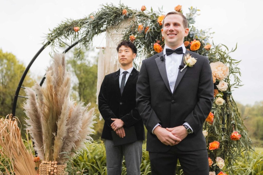 A groom in a black tuxedo and bow tie stands smiling beside a man in a black velvet blazer and gray trousers under a floral archway at the vineyard wedding ceremony.