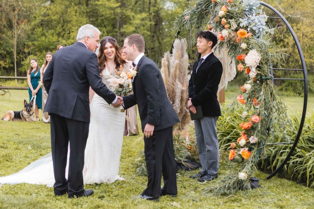 A bride and groom are at an outdoor wedding ceremony at The Barns at Hamilton Station, with the groom shaking hands with an older man. A floral arch is in the background, and guests and a dog are visible in the distance, adding charm to this vineyard setting.