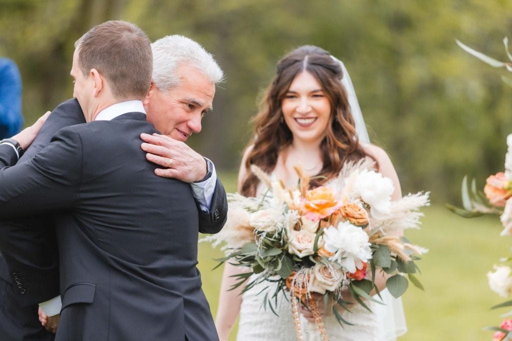 A bride holding a bouquet smiles while a grayhaired man and a younger man in suits hug outdoors at the wedding ceremony, with lush greenery in the background.