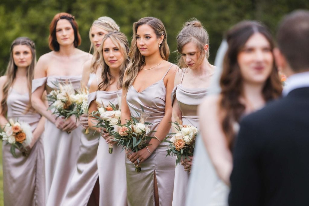 A group of bridesmaids in matching light silver dresses holding bouquets stand in a row during a picturesque vineyard wedding at The Barns at Hamilton Station, with the bride and groom slightly out of focus in the foreground.