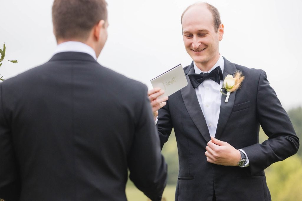 Two men in black suits stand facing each other outdoors at The Barns at Hamilton Station. One man, smiling, holds a card, possibly part of the wedding ceremony. The other man has his back to the camera. Both are dressed formally.