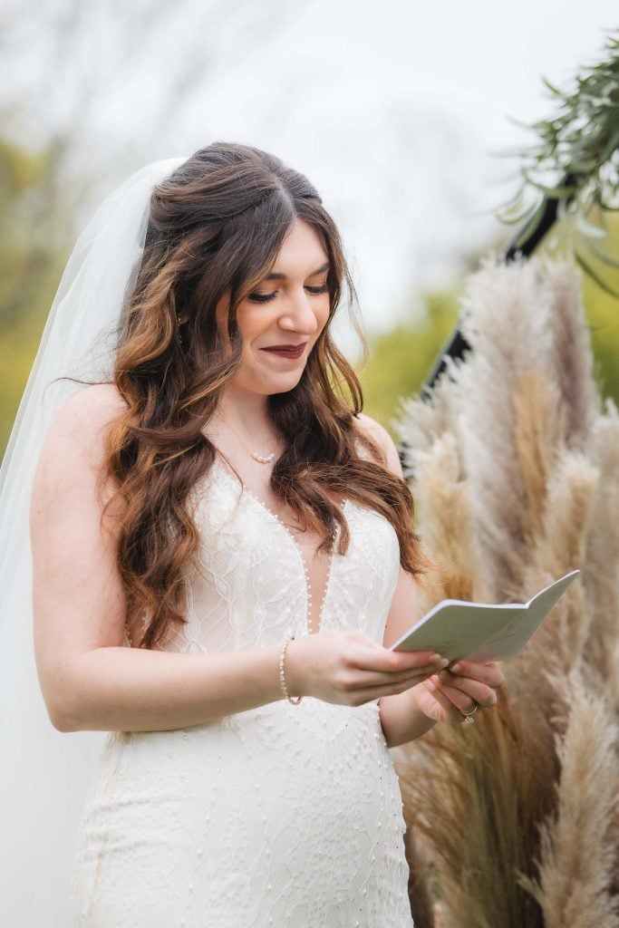 A bride in a white dress and veil stands outside, reading from a small booklet, with natural greenery in the background, setting the perfect scene for a vineyard wedding.