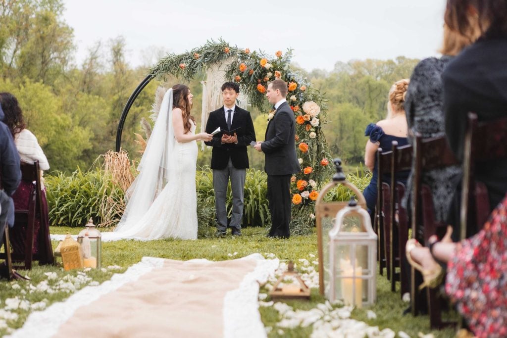 A bride and groom stand at an outdoor wedding altar at The Barns at Hamilton Station, officiated by a man. Guests are seated on both sides, and a floral archway is in the background, with the vineyard adding charm to the ceremony.