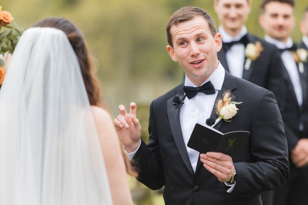 A groom in a black tuxedo is speaking while holding a booklet, facing a bride in a white dress and veil during an outdoor wedding ceremony at The Barns at Hamilton Station vineyard. Groomsmen are standing in the background.