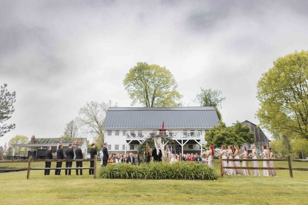 Outdoor wedding ceremony with guests seated facing a couple under an arch, in front of the rustic white building of The Barns at Hamilton Station, with a large tree and greenery in the background. Cloudy sky overhead.