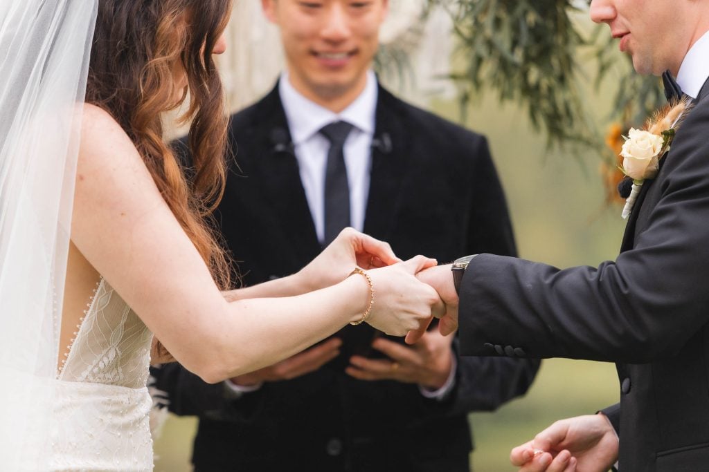 A bride and groom exchange rings during their wedding ceremony at The Barns at Hamilton Station, a picturesque venue nestled in a lush vineyard, with an officiant standing behind them.