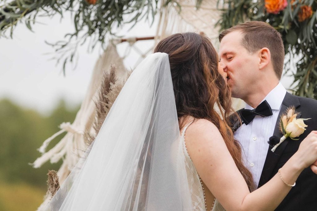 A bride and groom kiss during their outdoor wedding ceremony at The Barns at Hamilton Station, with greenery and floral decorations in the background.