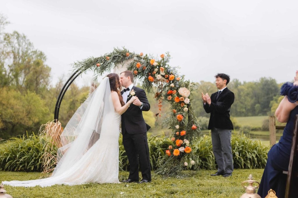 A bride and groom kiss under a floral arch during an outdoor wedding ceremony at The Barns at Hamilton Station, while a man stands nearby clapping.