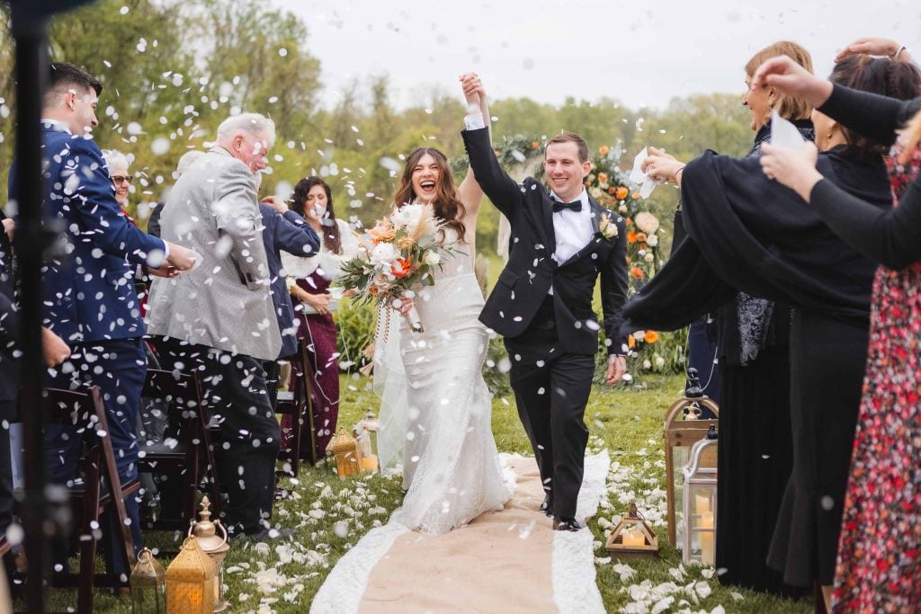 A newlywed couple walks down an outdoor aisle at The Barns at Hamilton Station, smiling and holding hands, as guests throw white confetti. The bride carries a bouquet and the groom wears a black suit. Lanterns and flowers decorate the picturesque vineyard setting.