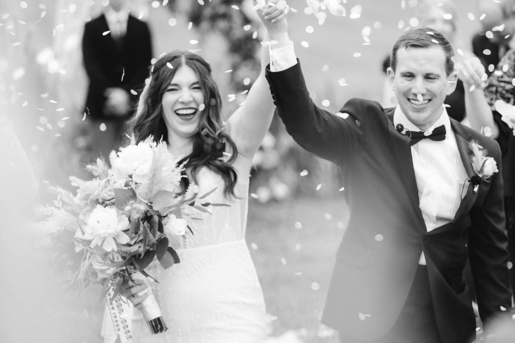 A bride and groom smile and raise their hands while walking down the aisle at The Barns at Hamilton Station, surrounded by flower petals. The bride holds a bouquet. The image, capturing the joy of their vineyard ceremony, is in black and white.