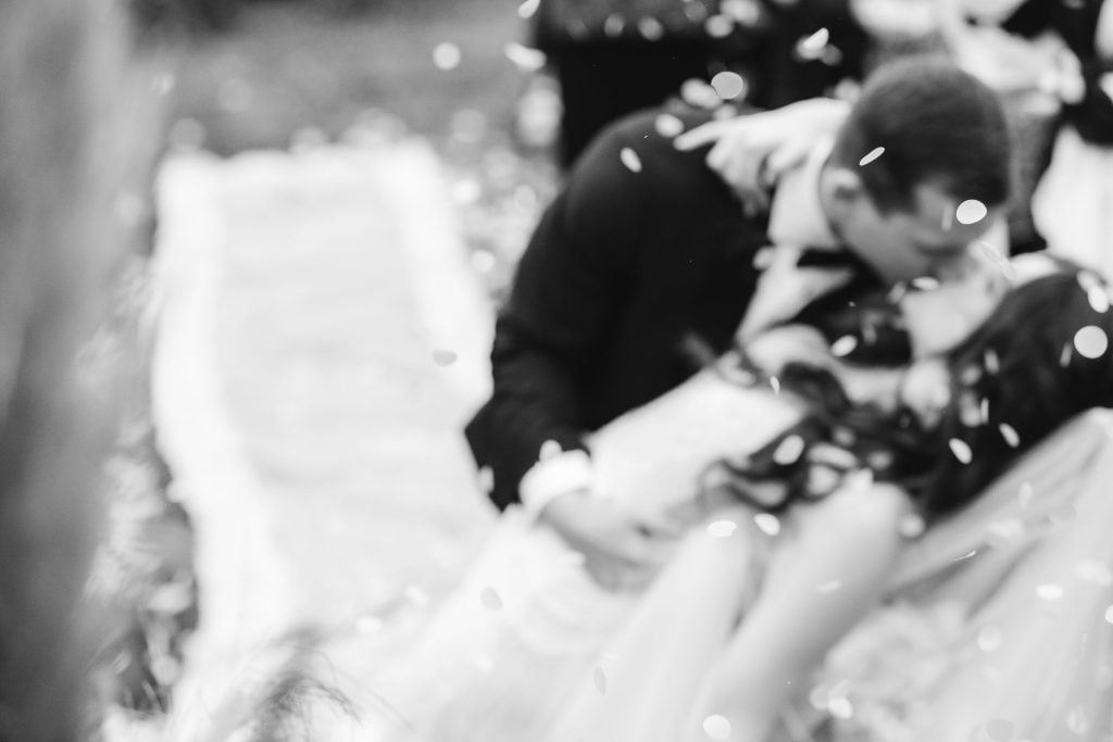 Black and white image of a bride and groom sharing a kiss during their ceremony at The Barns at Hamilton Station, while slightly blurry confetti floats around them. The groom leans over the bride as they embrace.