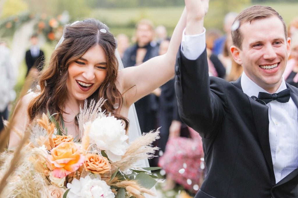 A happy couple, dressed in wedding attire, walks outdoors at The Barns at Hamilton Station while raising their arms in celebration. The bride is holding a bouquet of flowers, reflecting the joy of their ceremony amidst the picturesque vineyard backdrop.