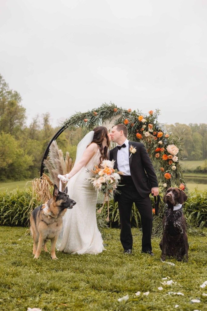 A bride and groom share a kiss under a flower arch at The Barns at Hamilton Station, with two dogs by their sides, adding a touch of charm to the vineyard wedding.