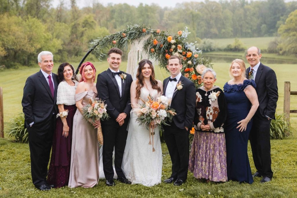 A wedding party stands outdoors, posing in front of a floral arch in a picturesque vineyard. The bride and groom are centered, surrounded by their family and friends, all dressed in formal attire.