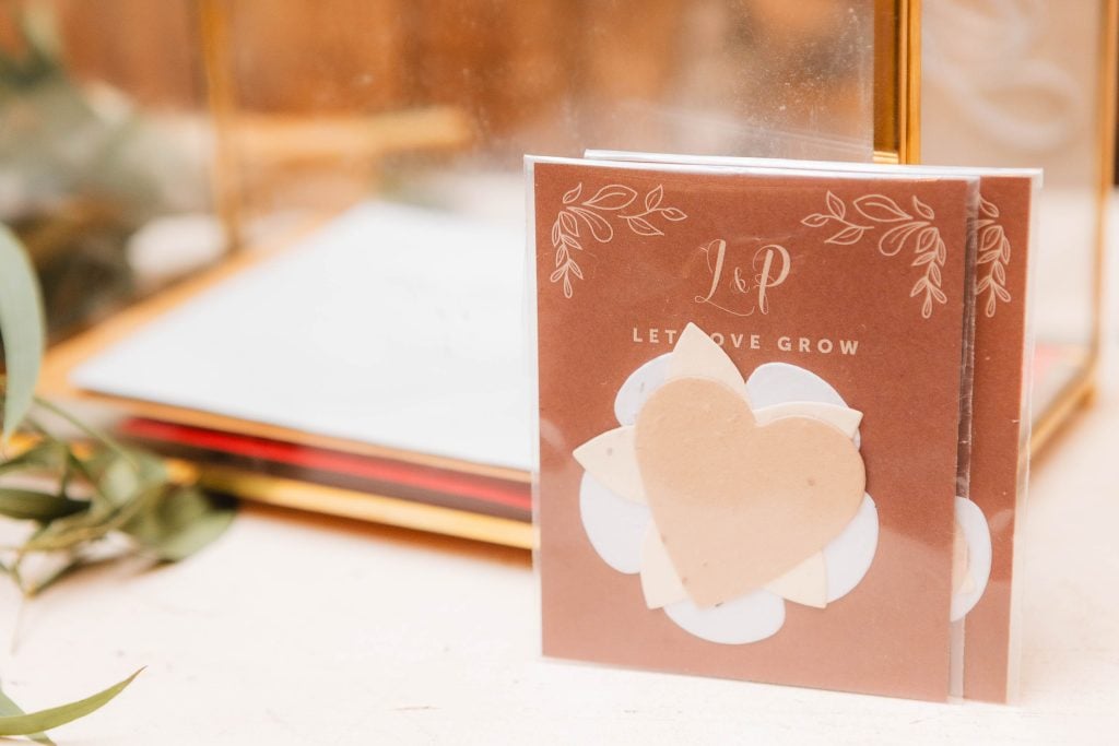 A small card with the text "Let Love Grow" and initials "L&P" is displayed, featuring a heart and leaf design. Set against a blurred background with golden accents, the intricate details perfectly complement a wedding at The Barns at Hamilton Station.