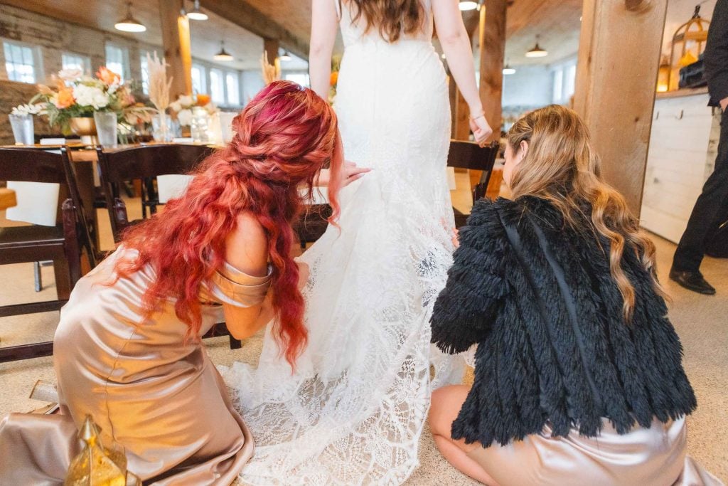 Two women in formal dresses adjust the lace train of a bride's white wedding gown inside The Barns at Hamilton Station, setting the perfect scene for an unforgettable wedding reception.