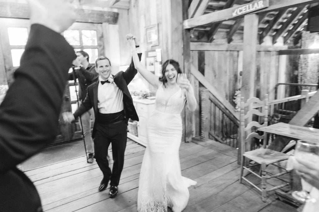 A bride and groom in wedding attire celebrate while holding hands and raising their arms at The Barns at Hamilton Station. Surrounding guests raise their hands in cheer during the rustic reception. The photo is in black and white.