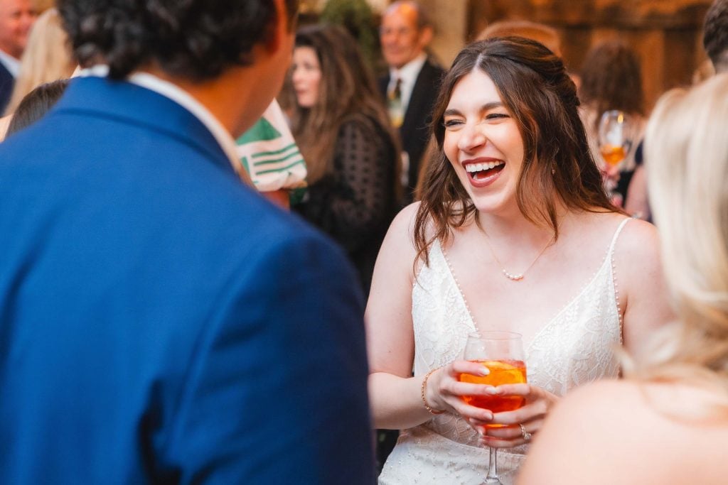 A woman in a white dress laughs while holding a drink at a wedding reception at The Barns at Hamilton Station, facing a person in a blue jacket.