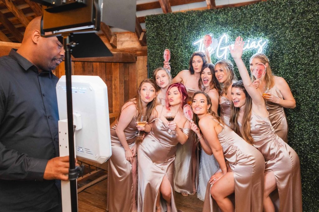 A group of women in lightcolored dresses pose for a photo with an attendant operating a photobooth at a wedding reception at The Barns at Hamilton Station, featuring a greenery backdrop and neon sign.