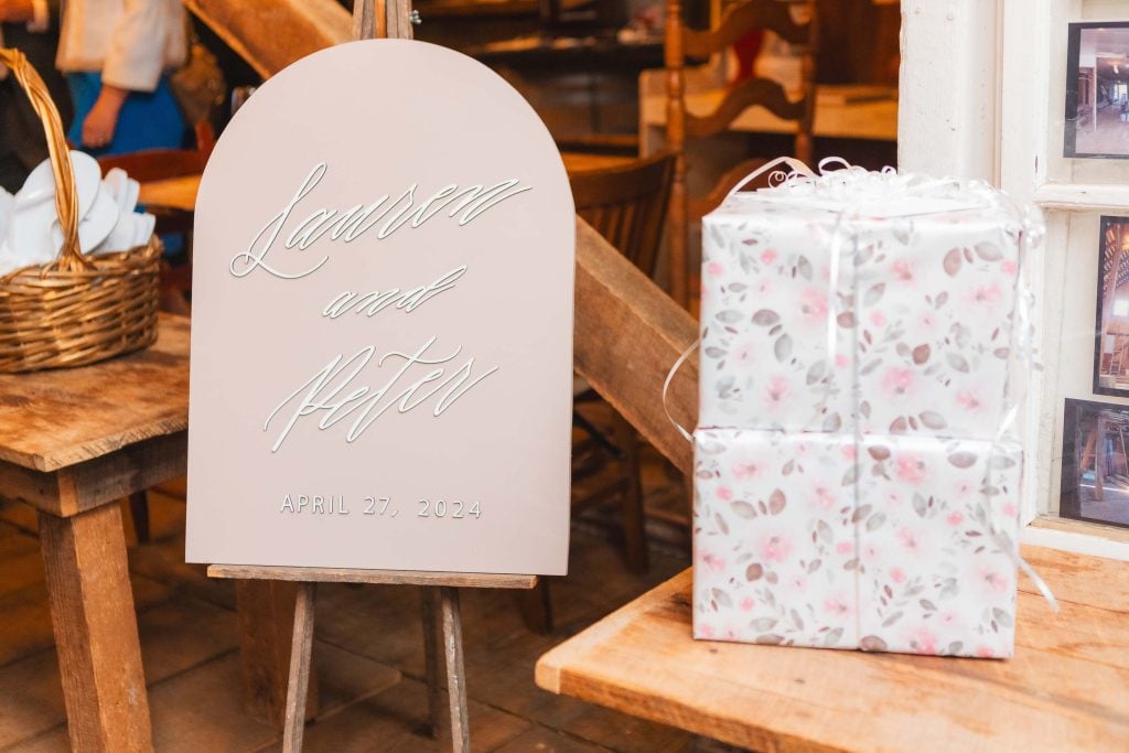 A wedding sign reads "Lauren and Peter, April 27, 2024" and is displayed next to a wrapped gift on a wooden table in the rustic setting of The Barns at Hamilton Station, capturing all the thoughtful details.