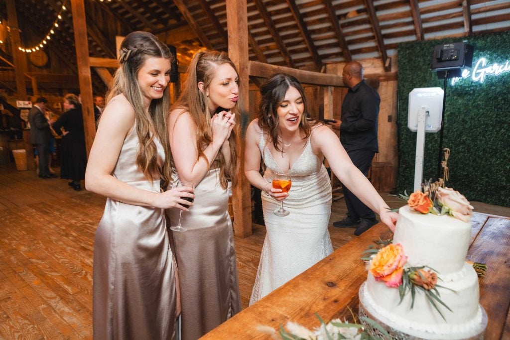 Three women in formal dresses stand by a decorated table with a tiered cake at The Barns at Hamilton Station. One woman in white reaches toward the cake, while the other two look on, one holding a drink. The charming woodpaneled room adds to the elegant wedding reception atmosphere.