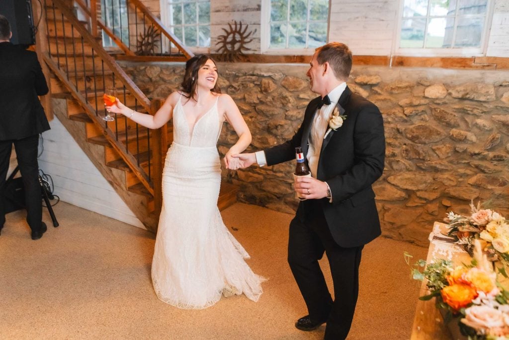 At The Barns at Hamilton Station, the bride in a white gown and groom in a black tuxedo dance while holding drinks, next to a wooden staircase in the rustic wedding reception setting.