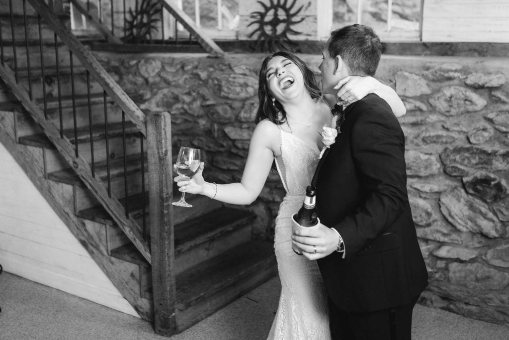 A bride and groom share a joyful moment, laughing and embracing, holding drinks in their hands beside a rustic stone staircase at their wedding reception at The Barns at Hamilton Station.