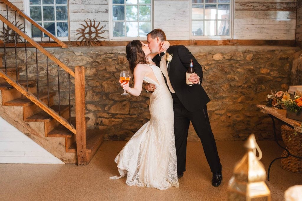 A bride and groom share a kiss while holding drinks in a rustic, stonewalled room with wooden stairs during their wedding reception at The Barns at Hamilton Station. Both are elegantly dressed, and the groom has a bottle in one hand.