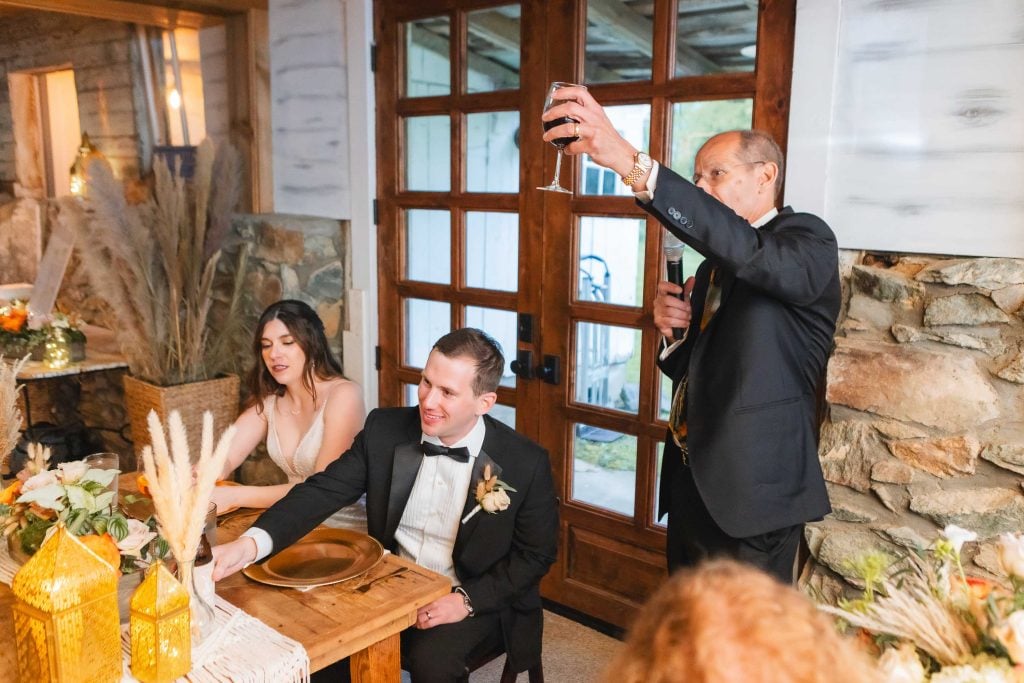 At the reception at The Barns at Hamilton Station, a man in a tuxedo raises a glass for a toast while standing next to a seated couple at a dining table. The woman is looking down, and the man is smiling.