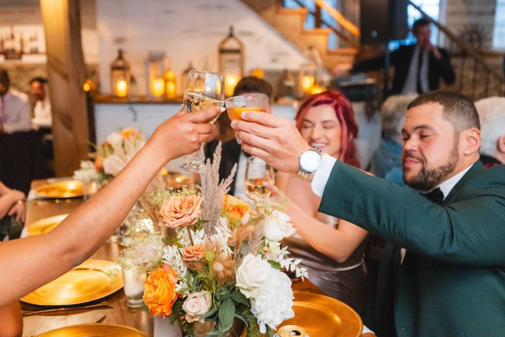 People clink glasses in a toast at a warmly lit reception at The Barns at Hamilton Station, the dinner table adorned with floral centerpieces and gold plates.