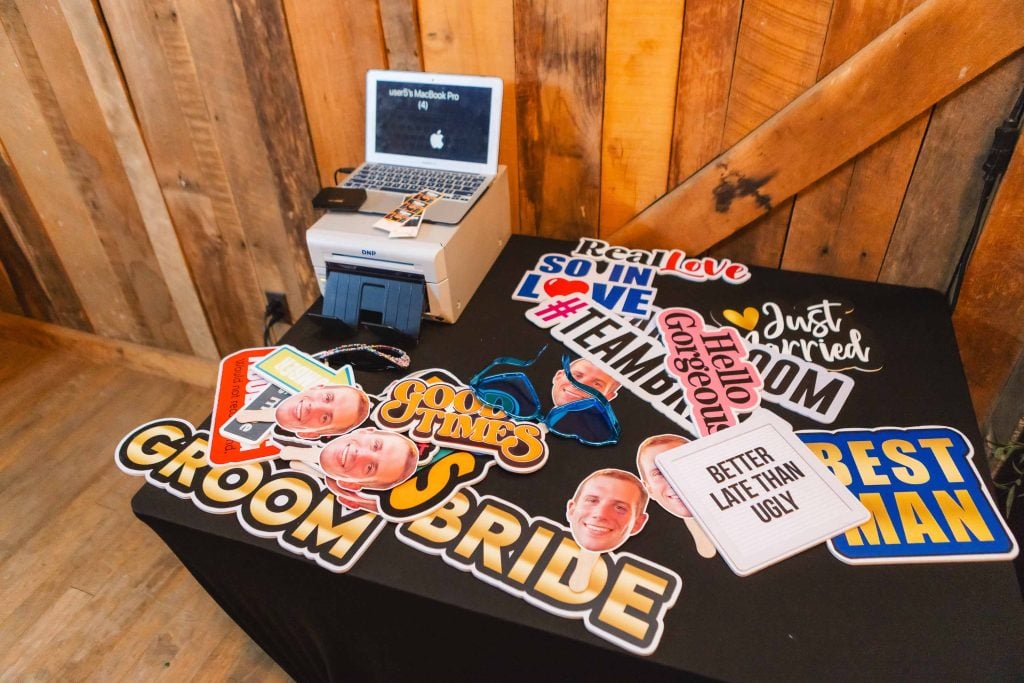 A table holds a laptop, a printer, and various photo booth props including text signs that say "Groom," "Bride," "Best Man," and other celebratory phrases at The Barns at Hamilton Station, showcasing charming details perfect for any rustic wedding.