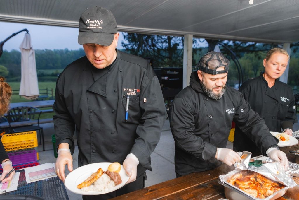 Three chefs wearing black uniforms prepare and serve food at an outdoor wedding reception. One chef is holding a plate with a meal, while the other two arrange food on serving trays at The Barns at Hamilton Station.