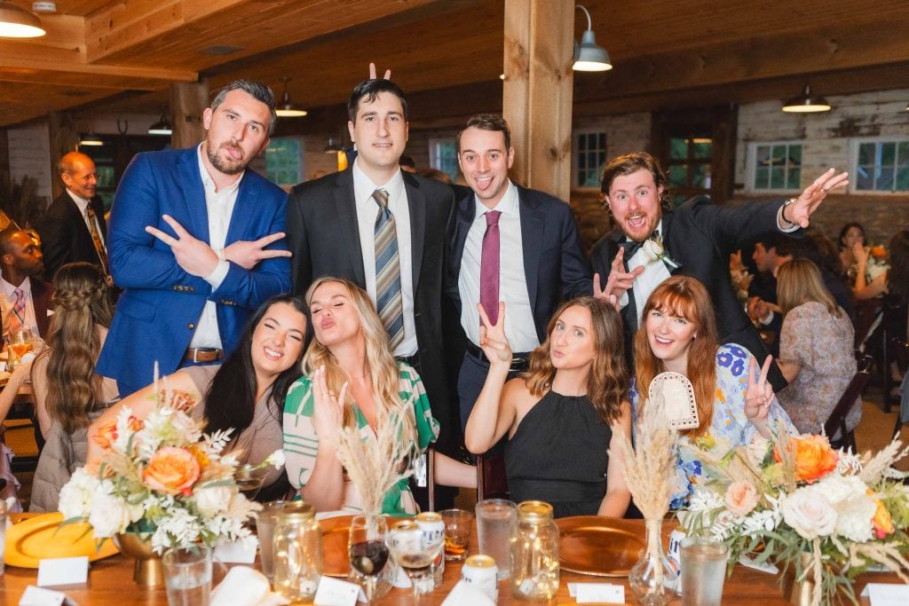 A group of eight people, formally dressed, pose together at a decorated wooden table during a wedding reception at The Barns at Hamilton Station. Some are making playful gestures with their hands, adding a touch of fun to the elegant setting.