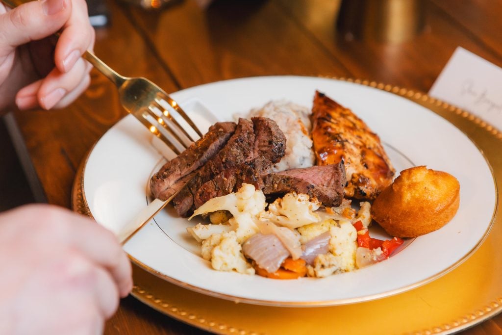 A plate with grilled meat, roast chicken, mashed potatoes, roasted cauliflower mixed with bell peppers and onions, and a cornbread muffin. A hand holding a fork and knife is visible on the left. The details of this feast were reminiscent of the sumptuous menus at weddings held at The Barns at Hamilton Station.