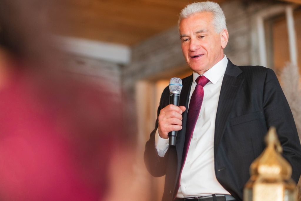 A man in a suit and tie speaks into a handheld microphone at the wedding reception held at The Barns at Hamilton Station.