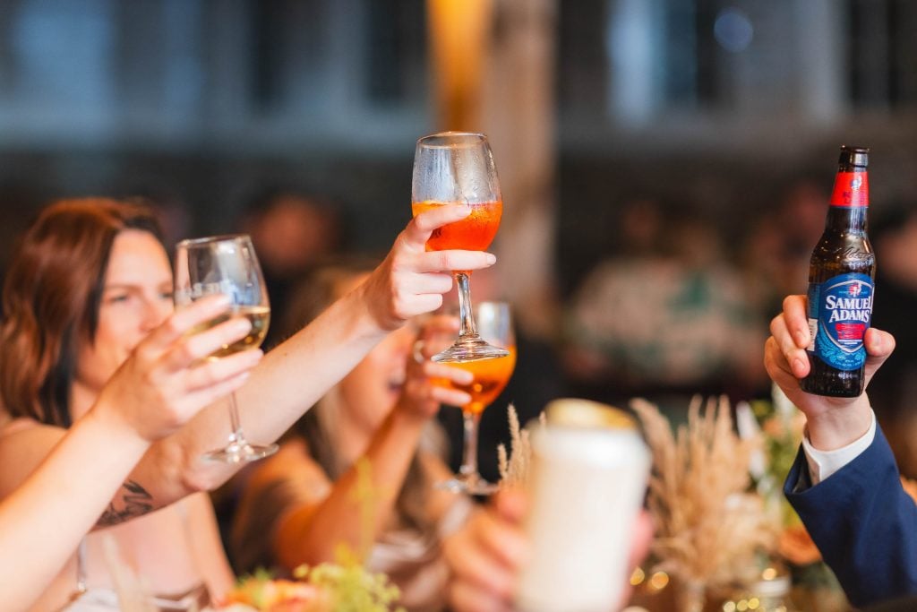 At a wedding reception at The Barns at Hamilton Station, people raise their glasses for a toast, holding various drinks including wine, beer, and a cocktail. The background is softly blurred.