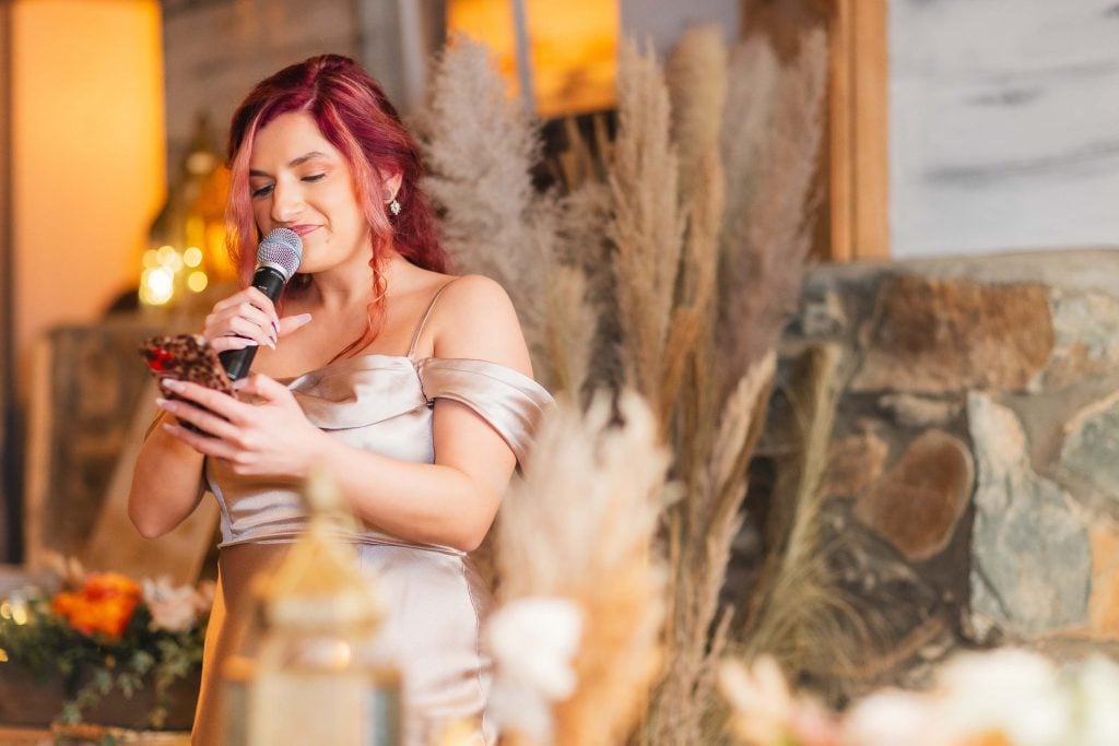 A woman with red hair is speaking into a microphone while holding a phone. She is wearing an offshoulder dress and standing in front of a rustic, decorated background with pampas grass and warm lighting, perfect for a wedding reception at The Barns at Hamilton Station.