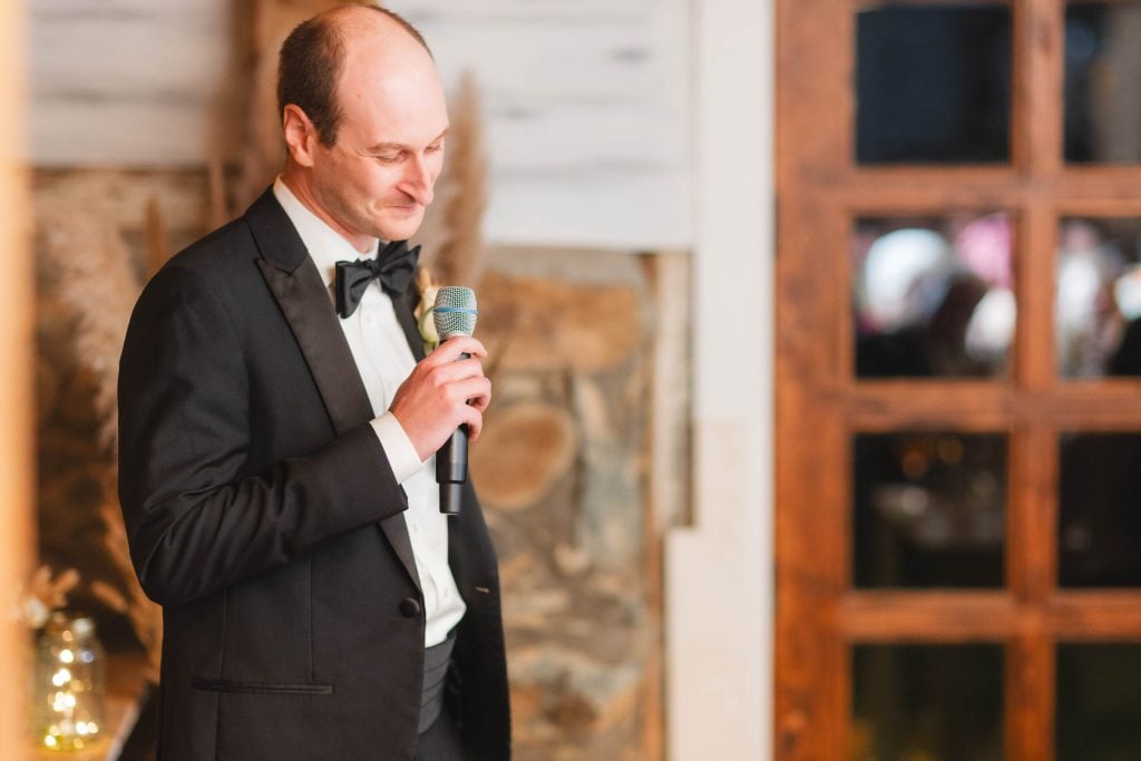 A man in a tuxedo holds a microphone, standing indoors near a wooden door and a stone wall, ready to make an announcement at the wedding reception held at The Barns at Hamilton Station.