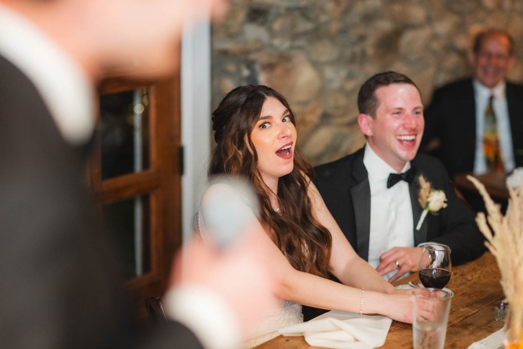 A bride and groom sit at a table, appearing amused and surprised, during a speech at their wedding reception at The Barns at Hamilton Station. A person with a microphone is in the foreground, blurred.