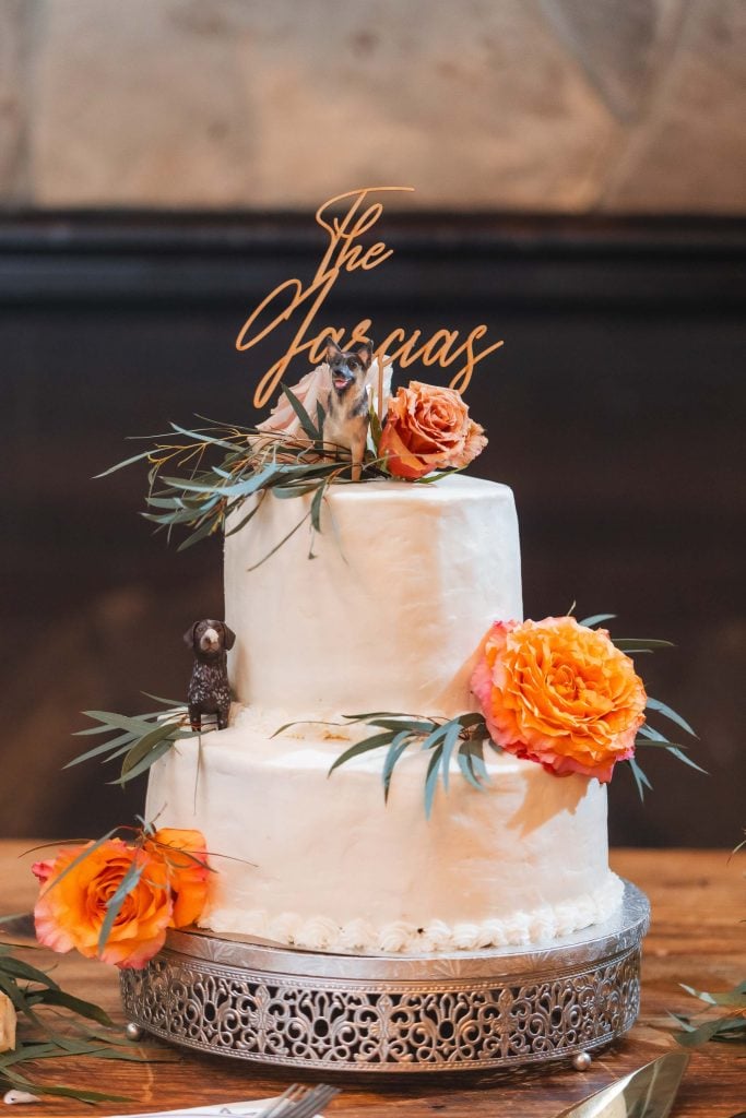 A twotier white wedding cake with orange flowers and leaf decorations, topped with figurines of dogs and a sign that reads "The Jaquas," perfectly complements the charm of The Barns at Hamilton Station. Every detail exudes elegance on this special wedding day.