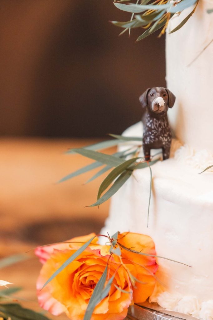 A small figurine of a brown and white dog stands on a white tiered wedding cake adorned with orange flowers and green leaves, capturing the charming details often seen at The Barns at Hamilton Station.