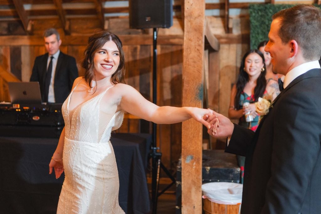 A bride and groom dance at their wedding reception at The Barns at Hamilton Station. The bride is smiling and holding the groom's hand as other guests watch in the warmly lit venue.