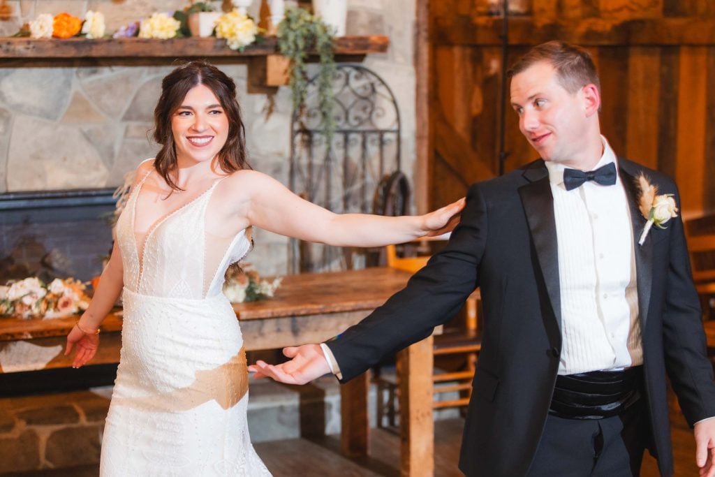 A bride in a white dress and a groom in a black tuxedo hold hands and smile at each other during their wedding reception at The Barns at Hamilton Station.