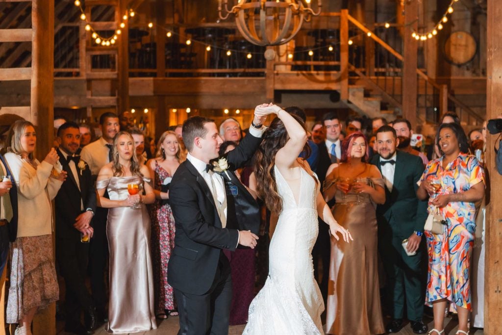 A bride and groom in formal attire dance at their wedding reception, surrounded by guests in a warmly lit, rustic venue at The Barns at Hamilton Station.