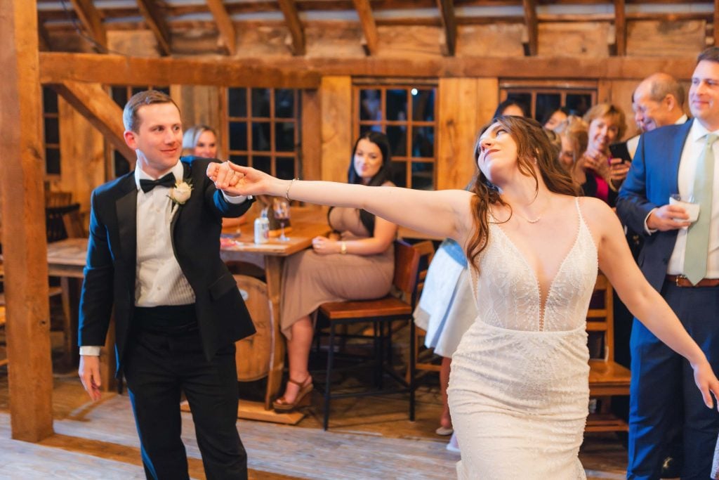 A couple in formal attire is dancing at a wedding reception at The Barns at Hamilton Station. The man is wearing a black tuxedo, and the woman is in a white sleeveless dress. Several people are seated and standing in the background.