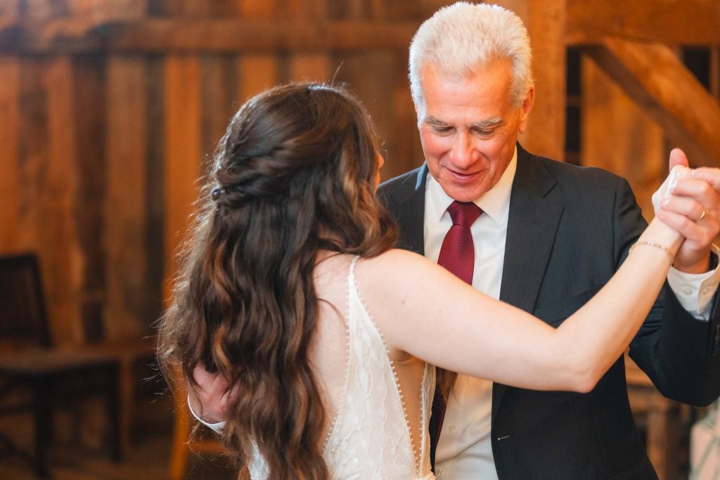 A woman with long dark hair in a white dress dances with a man in a black suit with a red tie. The backdrop of The Barns at Hamilton Station adds charm to their wedding reception, enhancing the rustic, wooden setting.