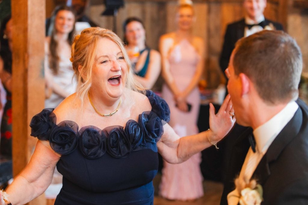 A woman in a dark blue dress is laughing and reaching out toward a man in formal attire at a wedding reception at The Barns at Hamilton Station, with other people standing and smiling in the background.