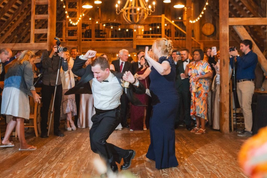 At a wedding reception in The Barns at Hamilton Station, a man in a suit and a woman in a dress dance energetically on the wooden dance floor. Guests around them watch, photograph, and cheer as the barn's rustic charm adds to the lively atmosphere.